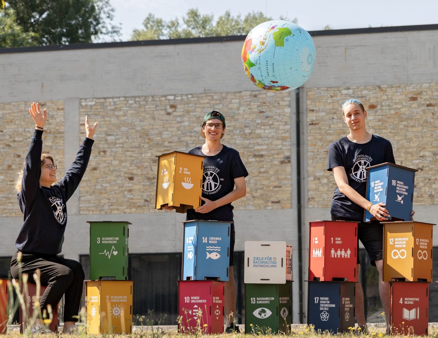Vier Studierende mit Hockern, auf denen die 17 Ziele für nachhaltige Entwicklung aufgedruckt sind. Zwei Studierende werfen sich einen Globus als Ball zu.